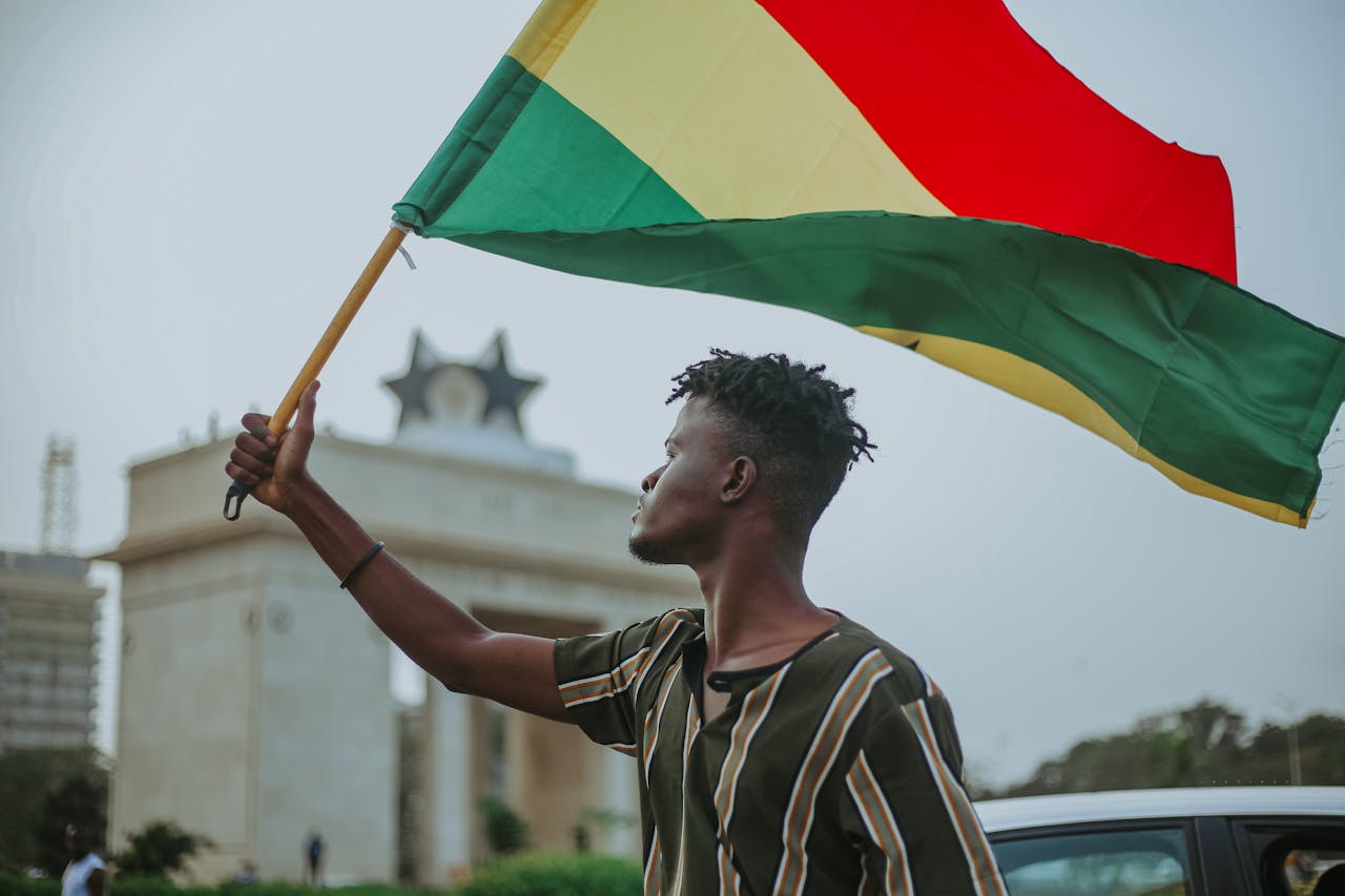 African male with dreadlocks raising flag of Ghana country with colorful stripes while looking away in town